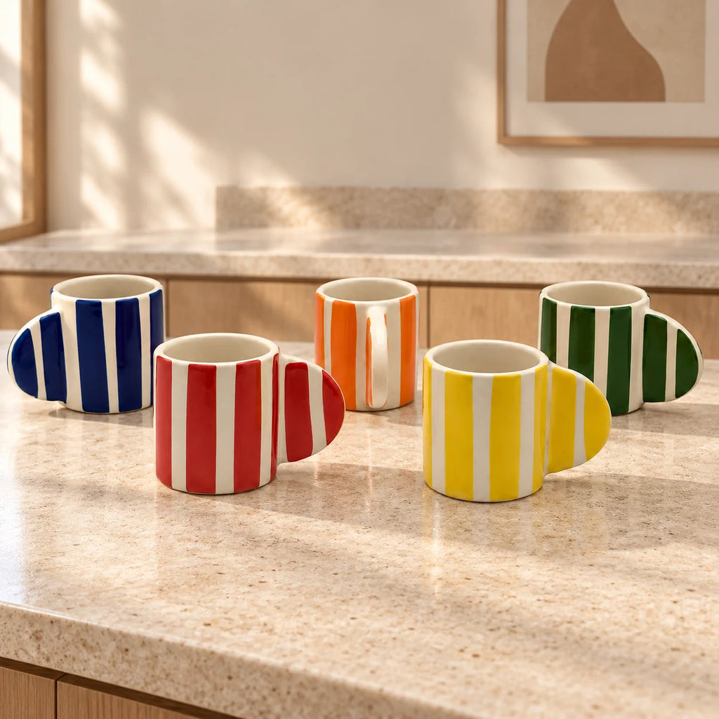 Five ceramic mugs with colorful vertical stripes and integrated handles are arranged on a beige stone countertop in a kitchen setting.