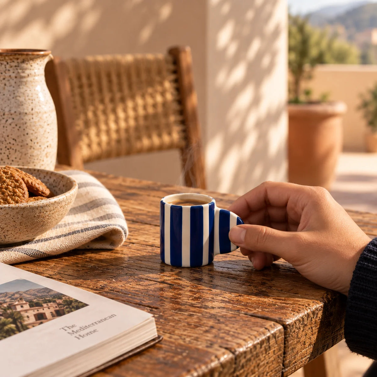 A hand holds a small ceramic espresso cup with bold blue and white vertical stripes on a rustic wooden table. The scene is styled with a book, a bowl of cookies, a woven chair, and a warm outdoor background.