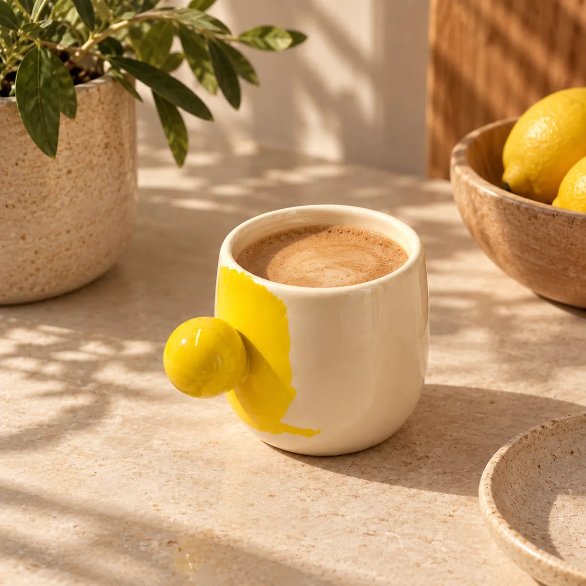 A white ceramic coffee mug with a protruding yellow lemon-shaped handle, filled with coffee, styled on a stone surface with a potted plant and lemons in the background.