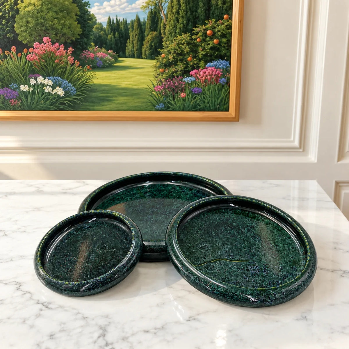 Three nested round trays in a dark green speckled stone-like glaze, displayed on a white marble surface with a framed garden painting in the background.