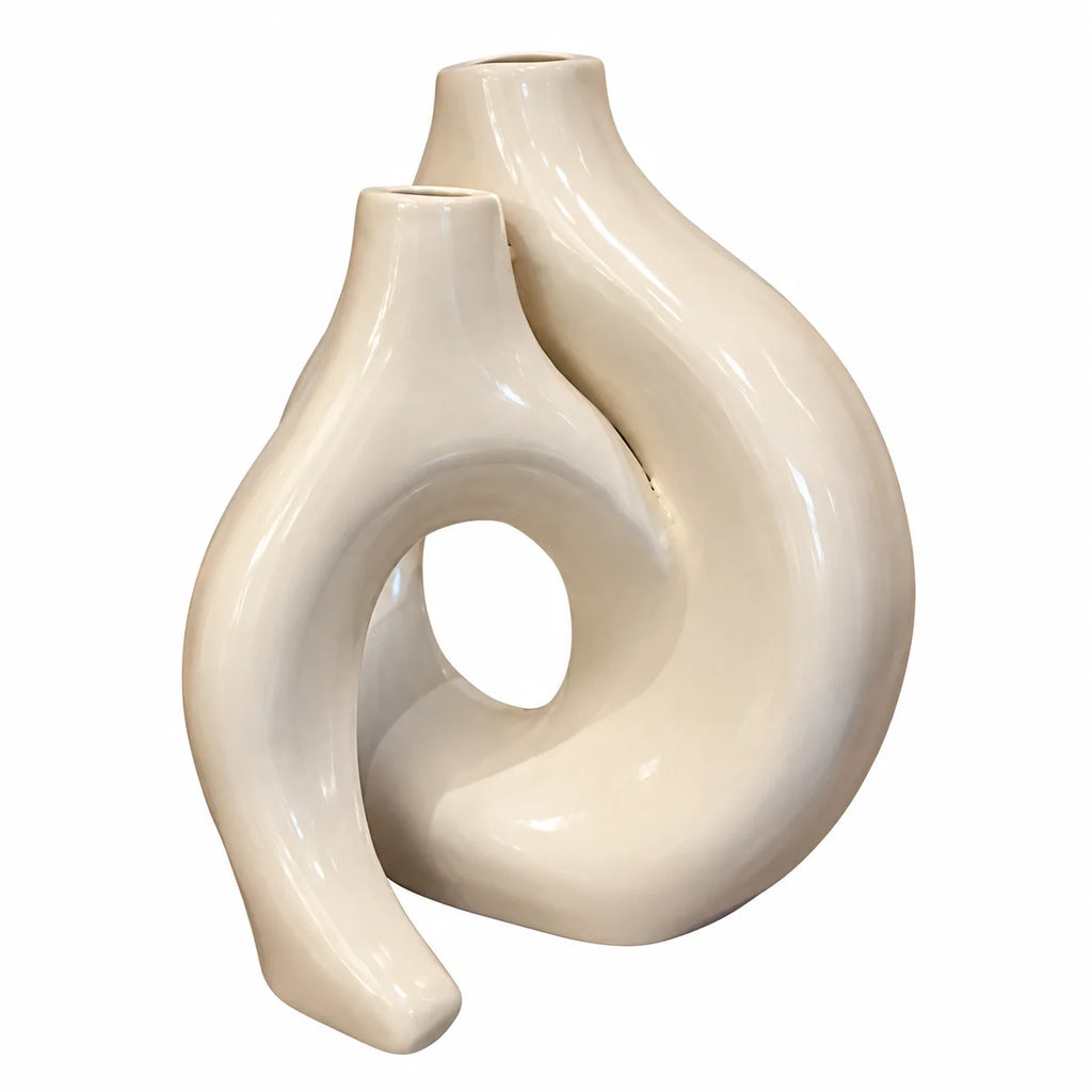 An abstract sculptural ceramic vase with a glossy finish, featuring two intertwined organic loops in a warm cream or off-white color, set against a plain white background.