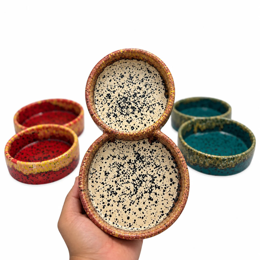 A hand holds a two-tier ceramic bowl set with a speckled beige interior and a speckled terracotta rim. Four additional matching bowls in red, yellow, teal, and green glazes are arranged in the background on a white studio background.