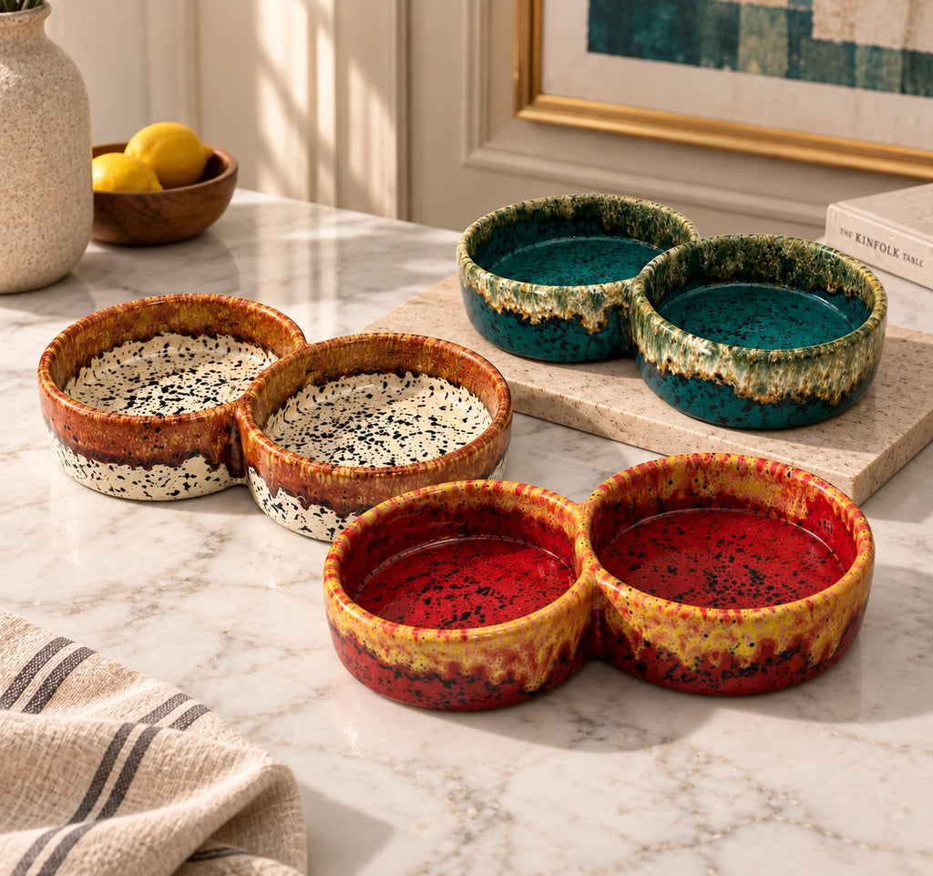 Four ceramic double-bowl coasters with speckled glazes in red, teal, and cream, arranged on a white marble countertop in a styled kitchen setting.